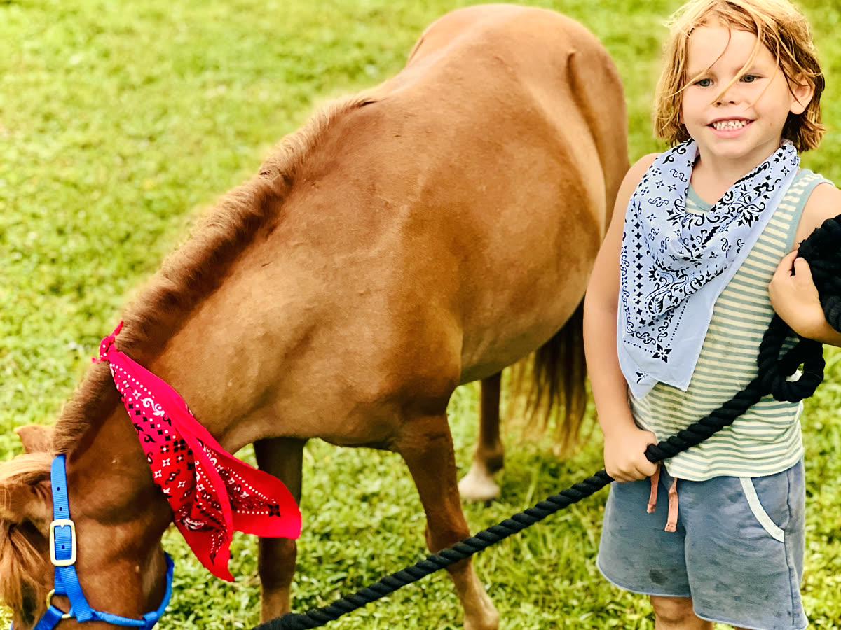 A boy holding a lead rope beside a chestnut pony wearing a red bandana